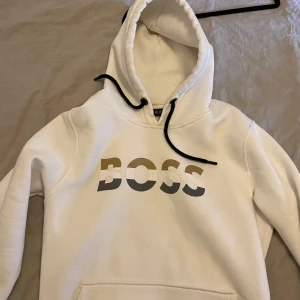Vit hoodie från BOSS - Vit hoodie från BOSS med svart snöre i huvan och stor logga framtill. Klassisk känguruficka och mjukt material, perfekt för en avslappnad stil.