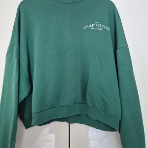 Grön sweatshirt från NLY Trend - Mörkgrön sweatshirt från NLY Trend med trycket 'Athletics Club' på bröstet. Tröjan har rund halsringning, långa ärmar och en något croppad passform. Perfekt för en avslappnad stil.