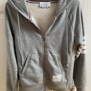 Thom Browne Hoodie - Säljer en grå hoodie från Thom Browne med dragkedja och huva. Tröjan har vita ränder på ena ärmen, fickor framtill och ribbade muddar. Klassisk och stilren design med märkets logga på bröstet.