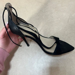 Svarta klackskor med snörning från Zara - Säljer ett par eleganta svarta klackskor från Zara med spetsig tå och tunna snören som går att knyta runt vristen. Skorna har hög klack och är tillverkade i syntetmaterial med mockakänsla. Perfekta för att lyfta din outfit.