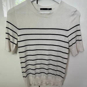 Randig vit tröja från 157, S - Vit stickad tröja från 157 med svarta horisontella ränder. Tröjan har rund halsringning, korta ärmar och en klassisk, enkel design som passar perfekt till jeans eller kjol. Mjuk och lätt i materialet för en chill look.