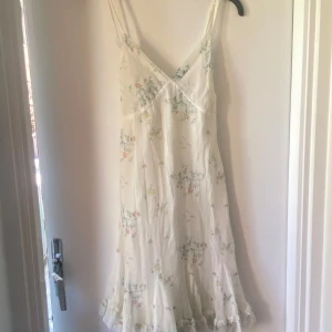 Vit blommig sommarklänning med volang - Superfin vit klänning med tunna axelband och blommigt mönster i pastellfärger. Klänningen har v-ringning, volangkant nertill och knytband i ryggen. Perfekt för varma dagar och har en lätt, luftig känsla.