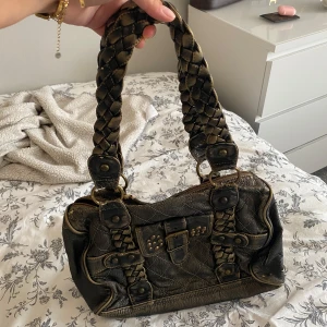 Vintage axelväska med flätade handtag - Cool vintage axelväska i skinnimitation med flätade handtag och snygga detaljer i form av nitar och spänne framtill. Väskan har dragkedja upptill, innerfack och dekorativa sömmar.