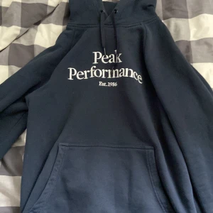 Mörkblå hoodie Peak Performance L - Snygg mörkblå hoodie från Peak Performance i storlek L. Klassisk modell med stor vit logga framtill, huva med snörning och känguruficka. Perfekt för chill dagar eller när du vill ha en clean och sportig look.