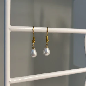 Guldiga örhängen med droppformad pärla - Snygga örhängen med guldfärgade krokar och droppformade vita pärlor. Perfekt accessoar för att lyfta din stil med en klassisk och elegant touch. Passar dig som gillar stilrena smycken med en twist. Handgjort 