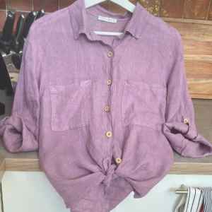 Ljuslila linneskjorta med knyt - Säljer en skjorta i 100% linne Skjortan har två stora bröstfickor, träknappar och ärmar som kan rullas upp och fästas med knapp. Den har en avslappnad passform och knytning framtill för en chill vibe. 100 % linne i färgen purpur/Syren. Lite rak i modellen och lite längre än en kort knytblus. Kan användas utan knyt med eftersom den är rak och vid nertill. Helt ny och endast provad! Säljer pga stor strl. Skulle säga att den passar 36- 42 beroende av om man vill ha lite oversize eller inte