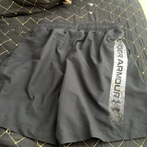 Svarta träningsshorts från Under Armour - Svarta träningsshorts från Under Armour med elastisk midja och lös passform. Snyggt tryck i silver längs ena benet med logga och text. Perfekta för gymmet eller löprundan, tillverkade i lätt och snabbtorkande syntetmaterial.