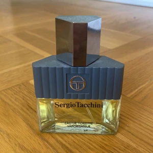 Sergio Tacchini Eau de Toilette - Stilren parfymflaska från Sergio Tacchini med geometrisk design. Flaskan är transparent med ljusgul vätska och har en grå, räfflad topp samt en silverfärgad, kantig kork. Perfekt för dig som gillar klassisk och modern stil.