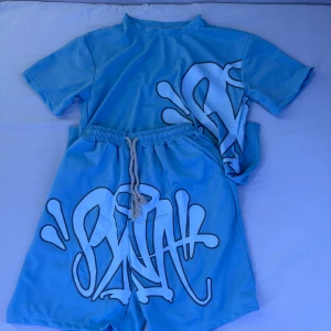 Blå t-shirt & Shorts från Synaworld (oanvända) - (Pris kan diskuteras vid snabb affär) Blå t-shirt från Syna World med stor svartvit graffiti-print över bröstet och ryggen. T-shirten har rund hals, korta ärmar och är gjord i ett mjukt material som känns skönt mot huden. Perfekt för dig som gillar streetwear och vill sticka ut.