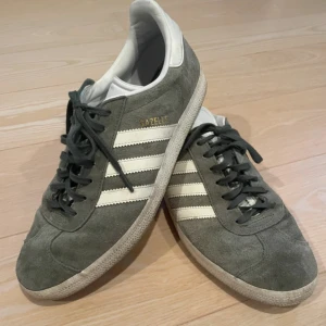 Adidas Gazelle gröna sneakers mocka - Klassiska Adidas Gazelle sneakers i grön mocka med vita detaljer och de ikoniska tre ränderna på sidorna. Skorna har svart snörning, vit plös och hälparti med Adidas-logga. Perfekta för en stilren och avslappnad look.