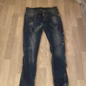 Mörkblå Dsquared2 jeans med slitningar - Säljer ett par mörkblå jeans från Dsquared2 med coola slitningar och patchdetaljer. Jeansen har skinny passform, klassiska fem fickor och snygga färgstänk. Perfekta för dig som gillar en edgy streetstil.