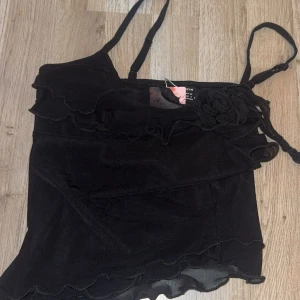 Svart volangig linnetopp från SHEIN - Supersöt svart linnetopp från SHEIN med tunna axelband, volangdetaljer och en dekorativ blomma framtill. Toppen har en lätt transparent känsla och är perfekt för dig som gillar trendiga och feminina plagg. Säljs med tag kvar!