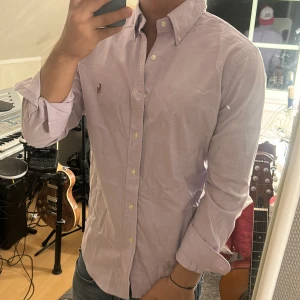 Ljuslila skjorta från Ralph Lauren - Säljer en stilren ljuslila skjorta från Ralph Lauren med klassisk button-down krage och broderad logga på bröstet. Skjortan har lång ärm, normal passform och är tillverkad i mjuk bomull. Perfekt för dig som gillar en clean och tidlös look.