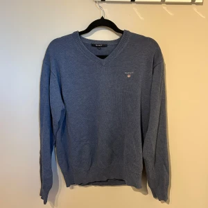 Blå stickad tröja från GANT - Klassisk blå stickad tröja från GANT med v-ringning och broderad logga på bröstet. Tröjan har ribbade muddar vid ärmslut och nederkant, perfekt för en stilren och avslappnad stil.