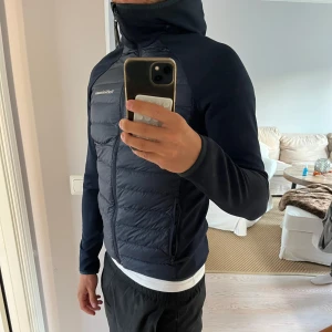 Peak performance cardigan  - Snygg mörkblå cardigan från Peak Performance med huva och dragkedja hela vägen. Jackan har quiltad framsida och släta ärmar, perfekt för kyliga dagar. Logga på bröstet och två dragkedjefickor. Sitter tajt och sportigt. Köpt för 1500kr, personen på bilden är 180!