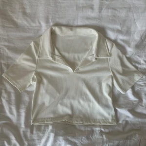 Vit ribbad topp med krage - Snygg vit ribbad topp med kort ärm och öppen krage. Croppad modell som ger en trendig look, perfekt till jeans eller kjol. Toppens ribbade material ger en extra touch och den har en stilren, enkel design