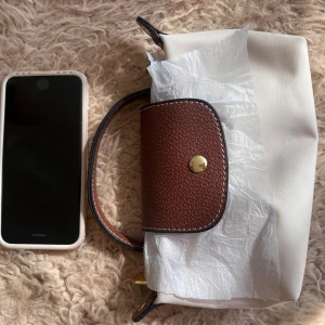 Beige och brun axelväska med detaljer lik Longchamp  - Snygg beige axelväska med bruna skinnhandtag och lock, guldfärgad knapp och dragkedja. Kommer med avtagbar axelrem i matchande brunt. Perfekt storlek för mobilen och det viktigaste. Stilren och enkel design som passar till många outfits.