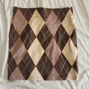 Brun rutig kortkjol från FB Sister - Superfin kortkjol från FB Sister i storlek XS med rutigt argylemönster i bruna och beige toner. Kjolen är figurnära och har en klassisk, preppy vibe. Perfekt att styla med en stickad tröja eller oversized t-shirt.