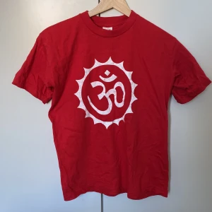Orange-Röd t-shirt med vit Om-symbol - Röd t-shirt från M&M i 100% bomull med en stor vit Om-symbol tryckt på bröstet. Klassisk passform och rund halsringning. Unik inköpt i Nepal, använd ett fåtal gånger 