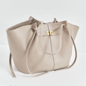 Gina Beige axelväska i skinn med gulddetalj - Gina Stilren beige axelväska i slätt skinn med snygga långa remmar och guldfärgat spänne framtill. Väskan har en rymlig form och minimalistisk design som gör den enkel att matcha med olika outfits. Perfekt för dig som vill ha en clean och modern look.
