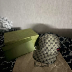 Gucci keps med GG-mönster - Exklusiv keps från Gucci i beige med klassiskt GG-monogram över hela kepsen. Kepsen har böjd skärm och en röd-grön rand på sidan som signaturdetalj. Tillverkad i canvas med läderdetaljer. Perfekt för dig som vill sticka ut med en lyxig accessoar.