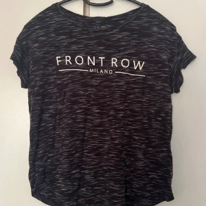 Svart mönstrad t-shirt Crocker XXS - Svart t-shirt från Crocker med vit text 'FRONT ROW MILANO' på bröstet. T-shirten har korta ärmar, rund hals och ett diskret mönster i vitt. Tillverkad i mjuk viskosblandning med polyester för extra komfort.