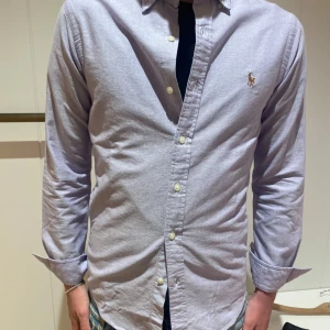 Ljusblå skjorta från Ralph Lauren - Säljer en stilren ljusblåskjorta från Ralph Lauren med klassisk button-down krage och broderad logga på bröstet. Skjortan har lång ärm, normal passform och är tillverkad i mjuk bomull. Perfekt för dig som gillar en clean och tidlös look. Med en liten fläck men inget man märker