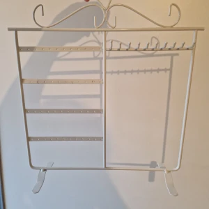 Vit smyckeshållare i metall - Snygg vit smyckeshållare i metall med romantiska detaljer upptill. Perfekt för att organisera örhängen, ringar och halsband. Flera rader med hål för örhängen och krokar för halsband eller armband. Stilren och passar lätt in i rummet.