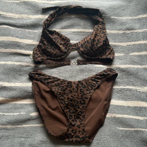 Brun leopardmönstrad bikini - Snygg bikini i brunt med svart leopardmönster. Överdelen har halterneck med breda band och bygel, samt transparent spänne i ryggen. Underdelen är klassisk med högre midja. Perfekt för dig som vill sticka ut på stranden!