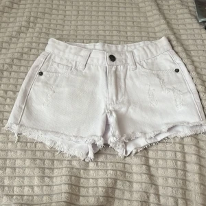 Vita jeansshorts - Snygga vita jeansshorts från UNIQ dnm (Ullared) med fransig  kant och slitna detaljer framtill. Klassisk femficksmodell med bälteshällor och knappstängning. Perfekta för varma dagar och en chill look. Storlek 134.