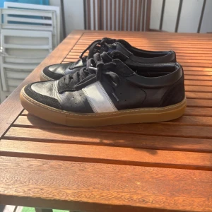 Svarta sneakers från Axel Arigato - Snygga svarta sneakers från Axel Arigato med vit rand på sidan och bruna gummisulor. Skorna har snörning, rund tå och är tillverkade i skinn och mocka. Perfekta för dig som gillar stilrena och sportiga vibes.