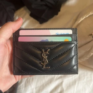 Svart YSL quiltad korthållare i skinn - Säljer en stilren svart korthållare från YSL i äkta skinn med quiltad design och ikoniska YSL-loggan i metall framtill. Perfekt för kort och ID, smidig att ha i fickan och ger en lyxig känsla varje dag.