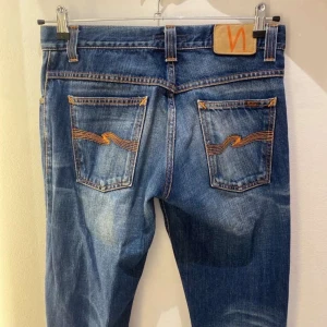 Blå jeans från Nudie Jeans - Säljer ett par klassiska blå jeans från Nudie Jeans med snygga slitningar och kontrastsömmar. Jeansen har raka ben, fem fickor och coola broderade bakfickor med Nudie-signaturen. Perfekt för dig som gillar en avslappnad men stilren look.