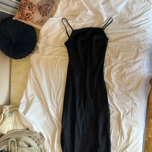 Svart långklänning med smala axelband - En stilren svart långklänning med smala justerbara axelband och rak halsringning, med dragkedja på sidan. Klänningen har en slät, glansig yta som ger en lyxig känsla och följer kroppen snyggt. Perfekt för dig som gillar minimalistisk och elegant stil.