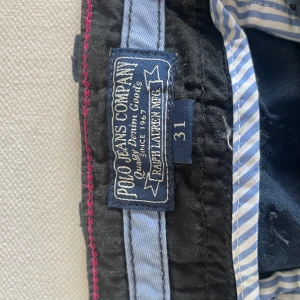 Mörkblå chinos från Polo Jeans Co - Mörkblå chinos från Polo Jeans Company Ralph Lauren i klassisk rak modell. Byxorna har sidfickor, knappdetaljer vid benslut och randigt innerfoder i midjan. Perfekta för en avslappnad men ändå stilren look.