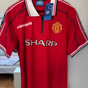 Manchester United Beckham 7 special edition  - Klassisk röd Manchester United fotbollströja från Umbro med SHARP som sponsor, klubbmärke på bröstet och vit-svart krage. På ryggen står det Beckham och nummer 7 med ett tryck av spelaren. Snygga detaljer längs ärmarna och dragkedja vid halsen. Den är ifrån 1999/00 säsongen. 