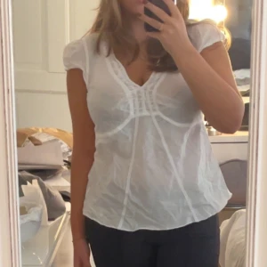 Vit blus med v-ringning och spetsdetaljer - Säljer en söt vit blus med v-ringning och fina spetsdetaljer framtill. Blusen har korta ärmar och lätt puff vid axlarna. Den är tillverkad i ett tunt, luftigt material som ger en romantisk vibe och passar perfekt till jeans eller kjol. Köpt secondhand men i bra skick! Passar strl M/L