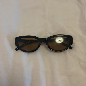 Svarta cat eye solglasögon - Snygga svarta solglasögon i cat eye-modell med bruna glas. Bågarna är i plast och har små guldfärgade detaljer vid gångjärnen. Perfekta för att lyfta din stil under soliga dagar.