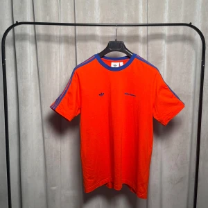 Wales Bonner X Adidas röd t-shirt, storlek M - En kultfavorit från ett av modevärldens mest omtalade samarbeten. Orangeröd (mer åt det röda) t-shirt med djupblå kontraster vid krage och ärmar. Tre klassiska ränder längs axlarna och tonade logotyper över bröstet.  Adidas till vänster, Wales Bonner till höger. Storlek M. Endast provad.