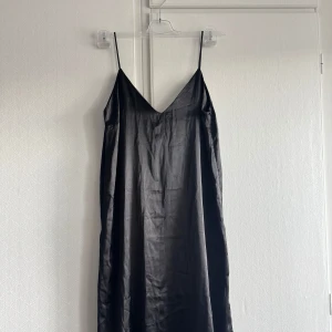 Svart långklänning från 157 Black Label - Svart långklänning i satinliknande material från 157 Black Label Collection. Klänningen har smala axelband och v-ringning framtill, vilket ger en stilren och elegant look. Perfekt för dig som gillar minimalistisk stil och vill ha något klassiskt i garderoben. Helt ny utan prislapp. 