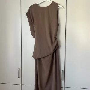 Brun långklänning med öppen rygg - Snygg brun långklänning med asymmetrisk design och cool öppen rygg. Klänningen har korta ärmar och ett twistat parti i ryggen som ger en unik look. Perfekt för dig som gillar stilrena och trendiga plagg med det lilla extra.