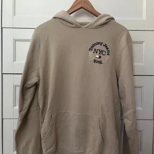Beige hoodie från Tommy Jeans XS - Beige hoodie från Tommy Jeans med broderad logga och texten 'Tommy Jeans NYC New York City' på bröstet. Klassisk känguruficka framtill, huva och lång ärm. Mjuk bomullskänsla och avslappnad passform, perfekt för chill dagar.