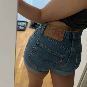 Levi's 501 högmidjade jeansshorts - Säljer ett par klassiska blå Levi's 501 jeansshorts med hög midja och uppvikta benslut. Shortsen har fem fickor, knäppning med knappar och den ikoniska läderpatchen bak. Perfekta för sommaren och ger en snygg vintage vibe.