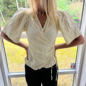 Vit omlottblus med puffärm - Superfin vit omlottblus med korta puffärmar och knyt i sidan. Blusen har diskret randigt mönster, v-ringning och spetsdetaljer längs kanterna. Materialet känns lätt och luftigt, perfekt för varmare dagar. Söt och trendig topp som lyfter vilken outfit som helst.