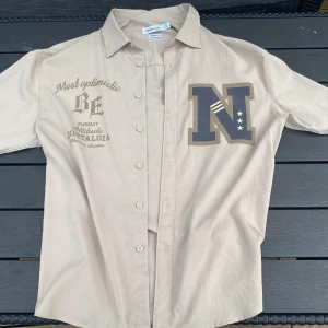 Beige skjorta från Yishion Casual M - Använd endast EN GÅNG, ca 22cm bred och ca 31 cm lång. LUFTIG och inte alls varm i sommarväder. Beige skjorta från Yishion Casual i comfort fit med tryck på bröstet och coola textdetaljer. Skjortan har klassisk krage, knäppning framtill och långa ärmar. Perfekt för dig som gillar en avslappnad men ändå snygg stil.