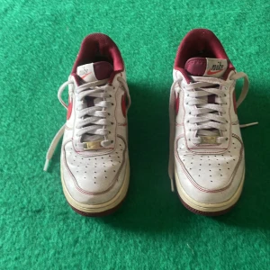 Nike Air Force 1 vit/röd sneakers - Säljer ett par klassiska Nike Air Force 1 sneakers i vitt med röda detaljer och swoosh. Skorna har röd insida, vita skosnören och perforerad tå för extra ventilation. Perfekt för dig som gillar retrovibbar och streetstyle.