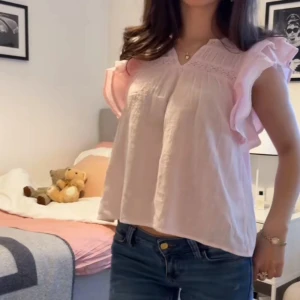 Ljusrosa blus med volangärmar - Supersöt ljusrosa blus med v-ringning och volangärmar. Blusen har broderade detaljer upptill och är tillverkad i ett lätt, luftigt bomullstyg. Perfekt för dig som gillar romantisk och feminin stil.