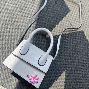 Vit axelväska från Jacquemus - Säljer en stilren vit axelväska från Jacquemus med krokopräglat mönster. Väskan har både handtag och lång axelrem, perfekt för att bära på olika sätt. Snygg och kompakt design med silverfärgad logga framtill.