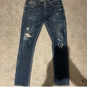 Dondup george med skönt slitage - Säljer ett par blå jeans från Dondup med coola slitningar och hål på benen. Jeansen har knäppning med knappar, klassiska fem fickor och orange sömmar på bakfickorna. Materialet är jeans i bomull och passformen är rak med normal midja.