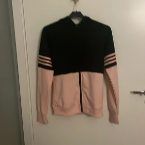 Svart och rosa Adidas tränningsjacka - Snygg trängningsjacka från Adidas i svart och ljusrosa med dragkedja framtill. Jackan har huva och tre klassiska ränder på ärmarna. Perfekt för dig som gillar sportig stil och vill sticka ut lite extra.